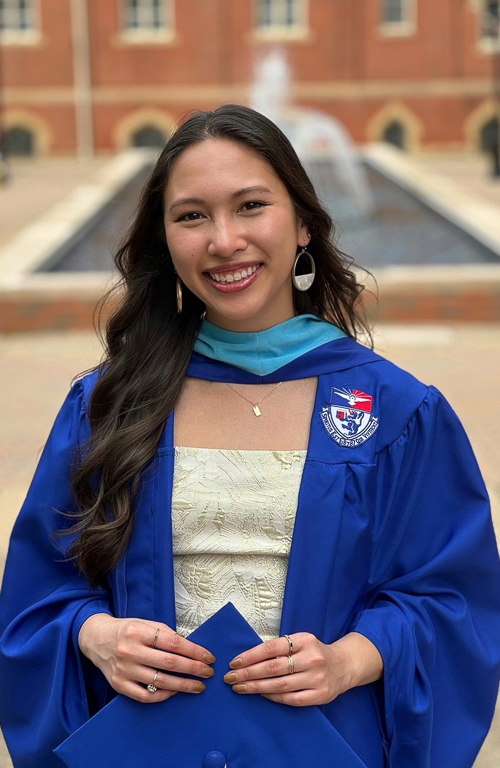 Photo of graduate Val Luutran in graduation cap and gown in front of water fountain on Duquesne campus