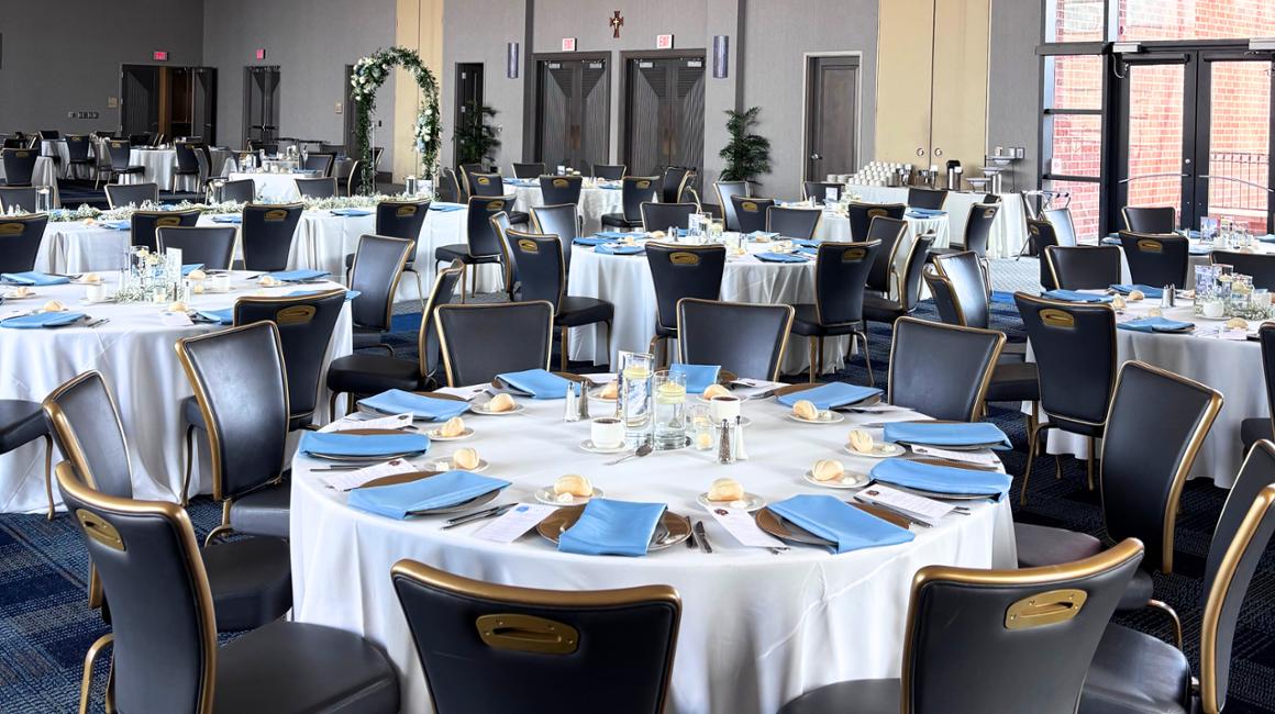 The Dougherty Ballroom, a large, well-lit banquet hall is arranged for a formal event. Multiple round tables covered with white tablecloths are evenly spaced throughout the room, each set with blue folded napkins, silverware, glassware, bread rolls, and small centerpieces. Dark padded chairs with gold trim surround each table. In the background, a decorative green and white floral arch stands near the wall. Along one side of the room are large windows and a beverage station with stacked cups and drink dispensers.