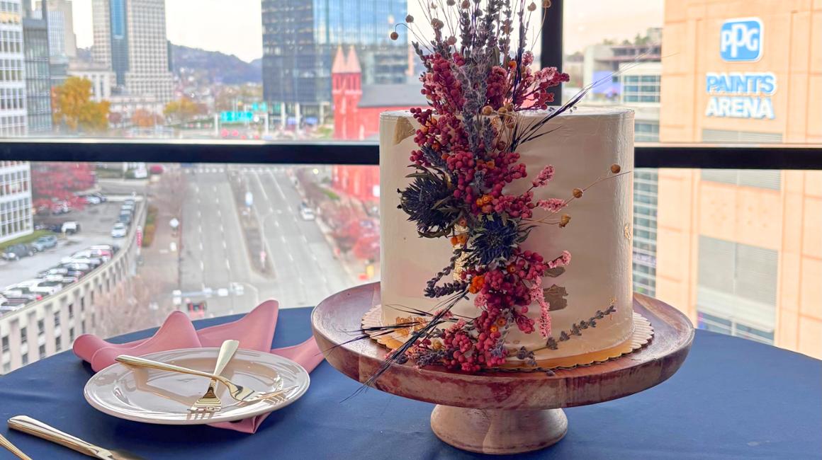 A single-tier round cake with smooth pale frosting sits on a wooden cake stand on a table draped with a dark cloth. The cake is decorated with an arrangement of dried flowers and berries in shades of pink, red, orange, and natural brown, cascading down one side. Next to the cake is a folded pink cloth napkin and a white plate with gold-handled fork and knife.