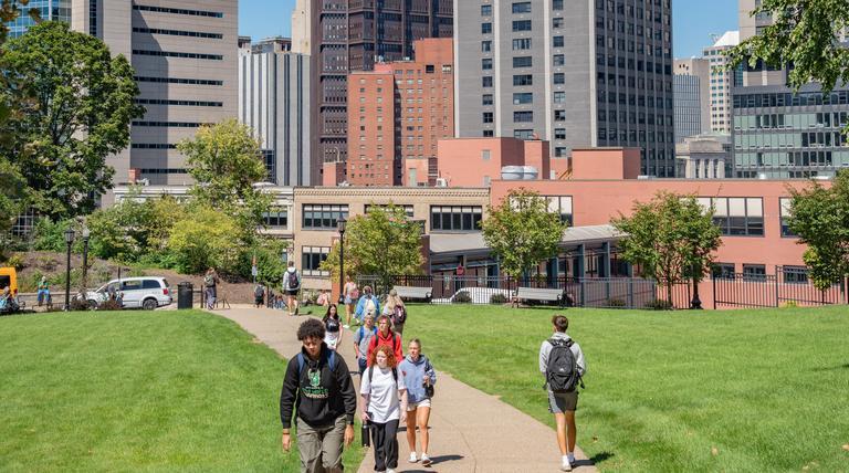 Duquesne students walking on green campus with city skyline in background