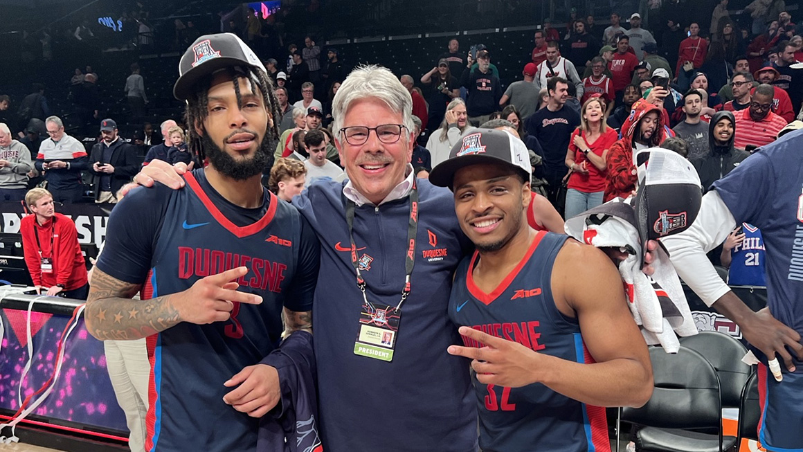 President posing with two Duquesne men's basketball players at the A10 championship game.