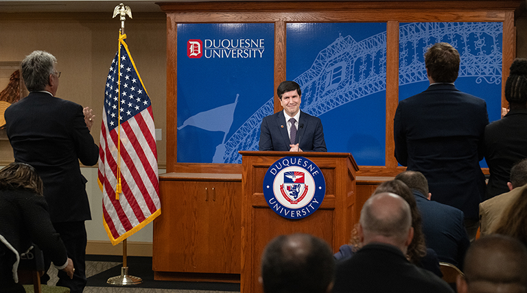 David Dausey speaking at a podium while people stand and applaud