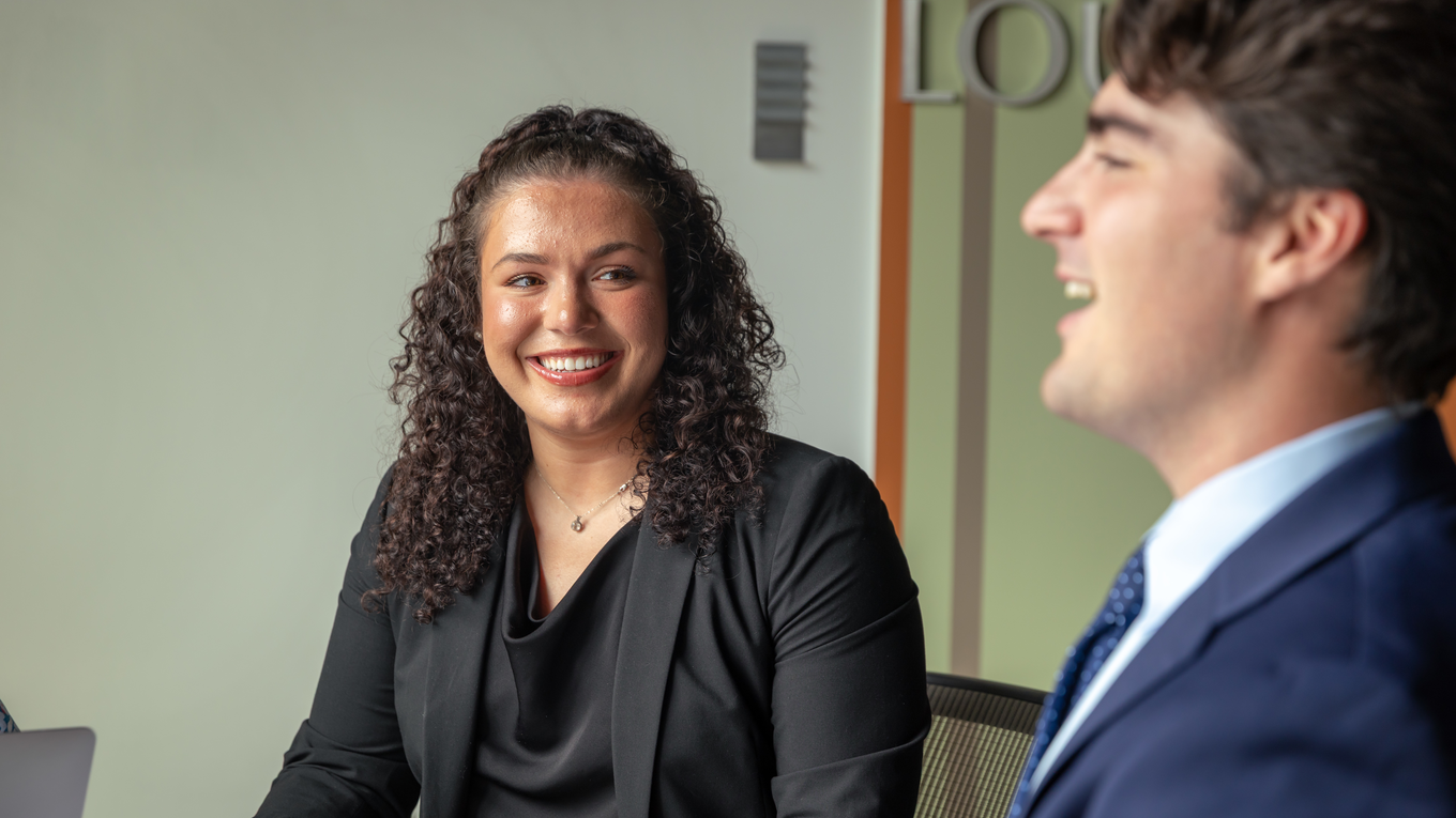 A student dressed in business professional attire smiles in a conference room.