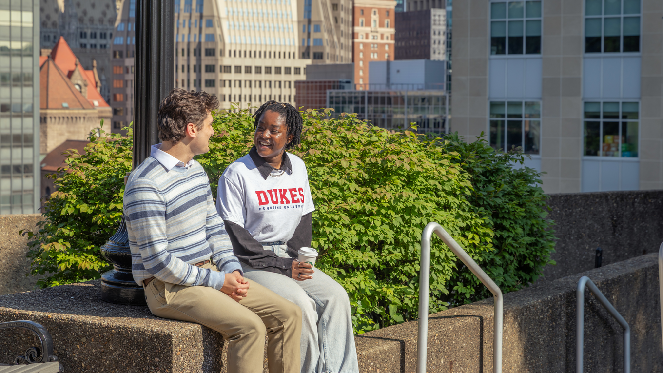 Two students chat outside on Duquesne's campus with the Pittsburgh skyline in the background.