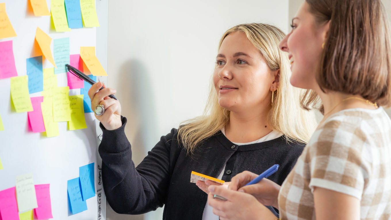 Two graduate students brainstorm on a whiteboard with colored sticky notes.