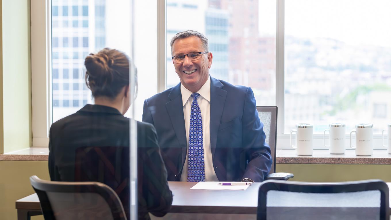 A business professional in a suit smiles at a colleauge. 