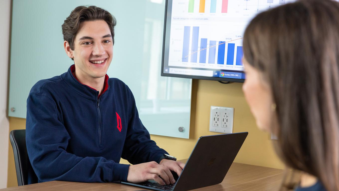 A student sits at a table and smiles. Bar graphs are on the screen behind him. 