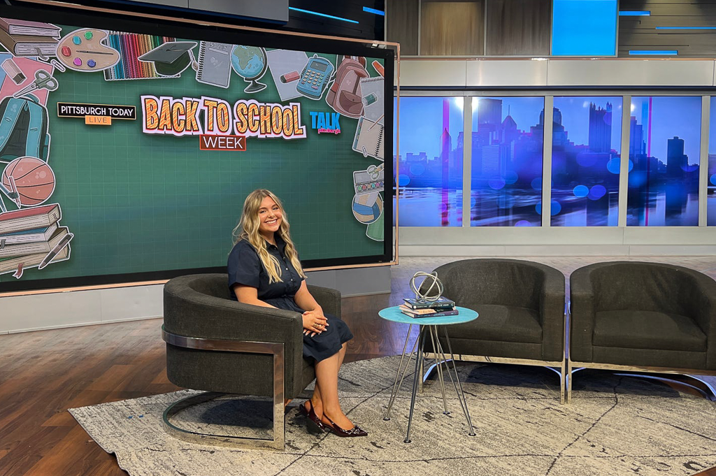 School of Education student Zoe Brunick on KDKA Talk Pittsburgh set for Teach Me Tuesday show seating in chair with table with books in front and backdrop of TV show titles reading Pittsburgh Today Live; Back to School Week; Talk Pittsburgh.