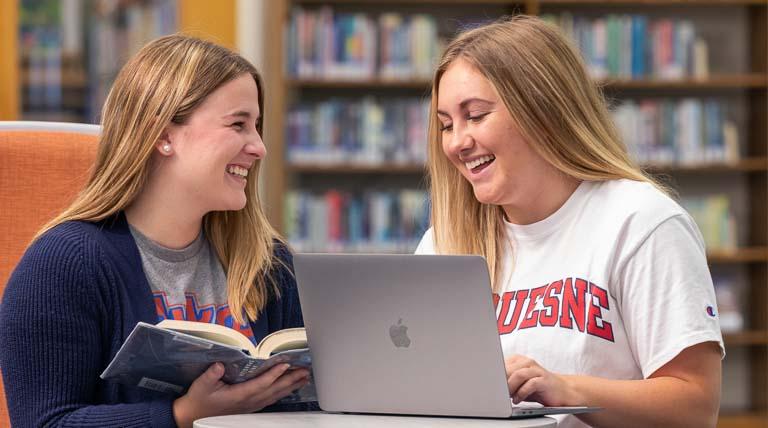 Dual degree students studying together in the library around laptop in chairs at small round table with colorful bookshelves in the background