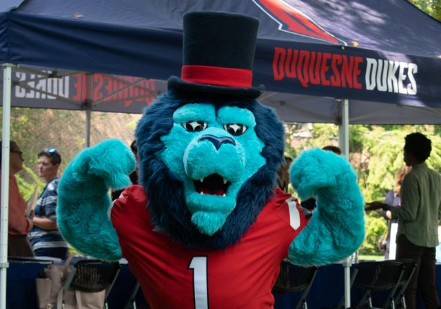 Duquesne Duke lion mascot flexing at outside campus event with tent in background.