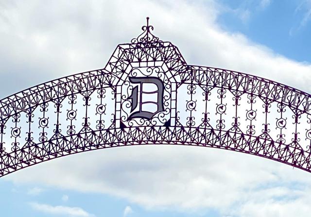 Duquesne University roadway arch of a metal rod curve arch structure with Duquesne D in center set against background of clouds and blue sky.