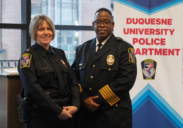 Two Duquesne public safety officers in uniform with sign in background that reads Duquesne University Police Department with Duquesne police badge logo on sign