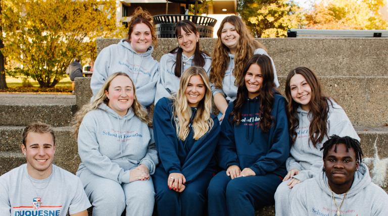 Nine School of Education Scholarship students gathered sitting on cement stadium steps outside on fall day.