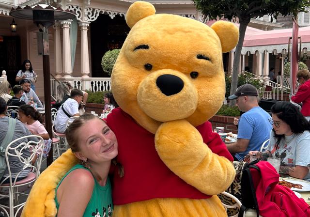 School of Education Dual Certification student Phoebe McNeil smiling at cafe table outside as life-sized Winnie-the-Pooh character hugs her at Disney.