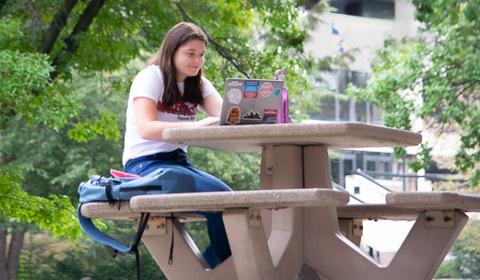 School Psychology student studying outside on campus on laptop at table with tres and campus building in the background