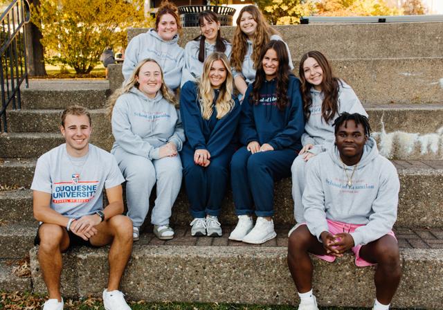 Nine School of Education Ambassadors smiling seated in group on stadium steps outside with fall trees in background