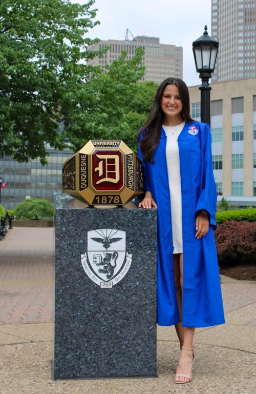 B.S.Ed. Secondary Education alum in graduate regalia on campus by Duquesne ring monument and green trees and city buildings in background