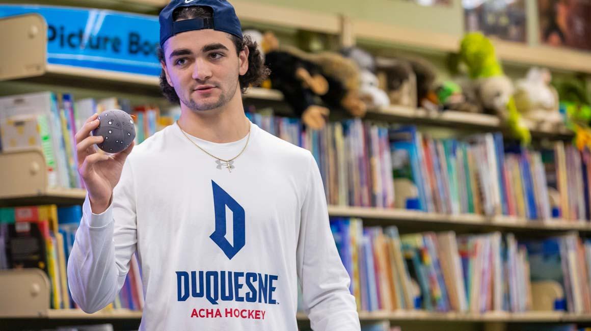 B.S.Ed. Secondary Education student holding ball wearing Duquesne hockey shirt doing mock lesson in the library curriculum center with colorful books and puppets behind him