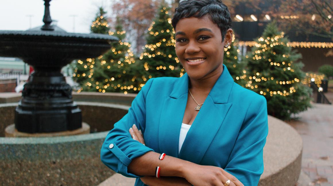 Fulbright Scholar and Ph.D. School Psychology student outside union smiling with arms crossed wearing blazer and Christmas trees with lights in background