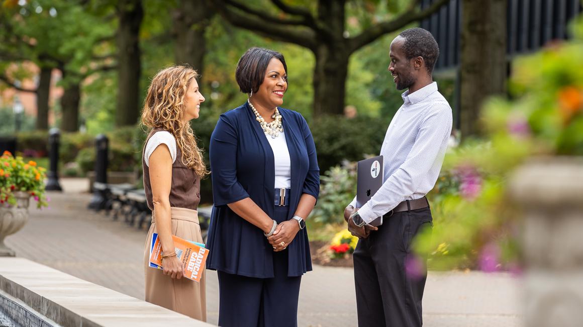 Photo of Dean with student and faculty member on Academic Walk outside Canevin Hall conversing in early fall with flowers in bloom