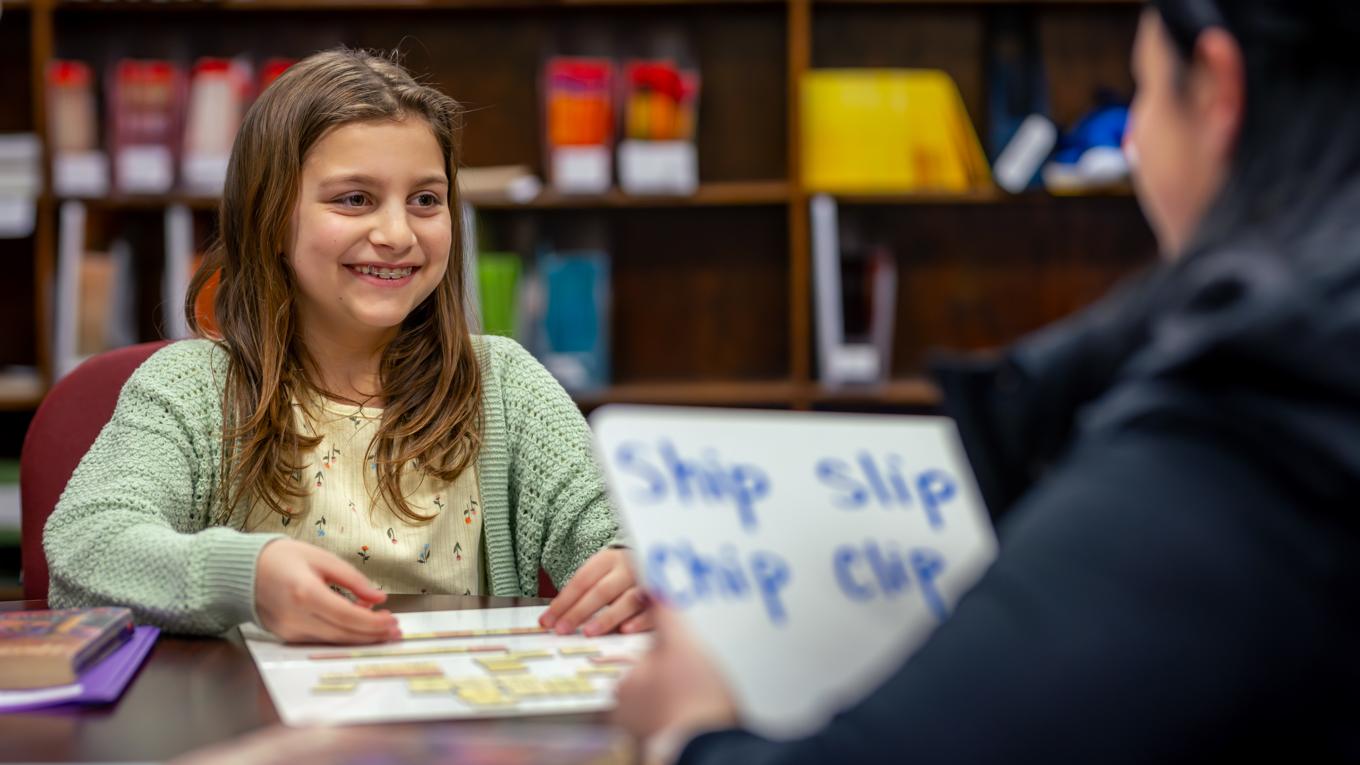 M.S.Ed. Reading and Literacy Education student engaging in reading lesson with white board with words ship slip chip clip with young student at table with bookshelves in background.