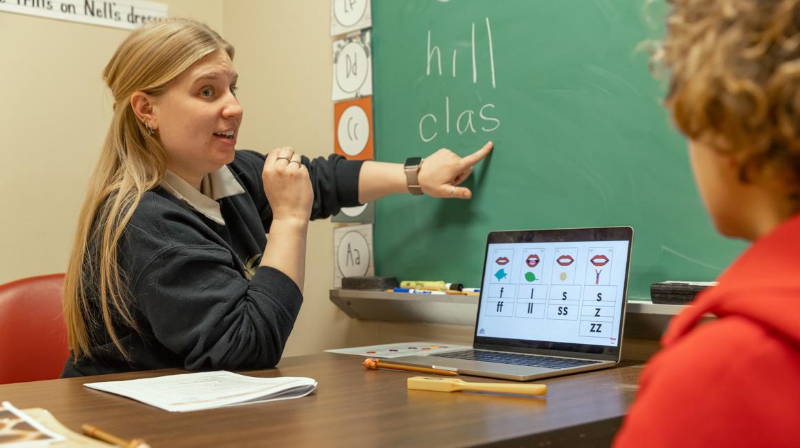 Reading clinic instructor with student engaging in reading lesson point to board with hill and clas written and laptop on desk and papers and school supplies between them. 