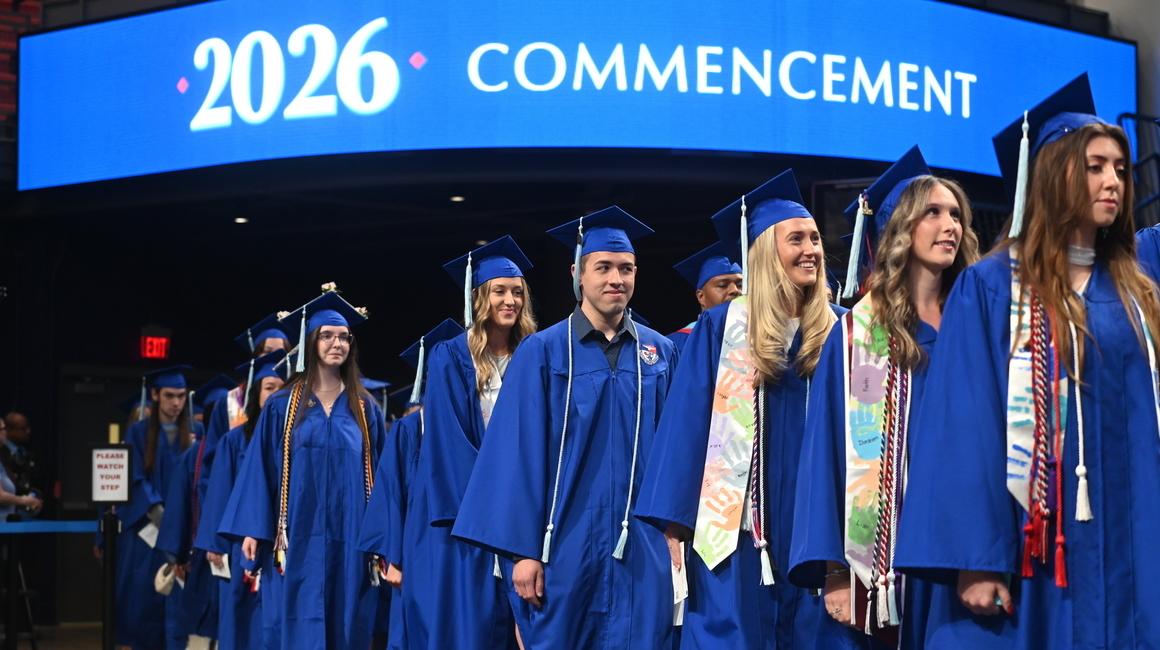 Line-up of spring School of Education new alumni dressed in regalia on commencement floor digital board above reading 2026 Commencement.