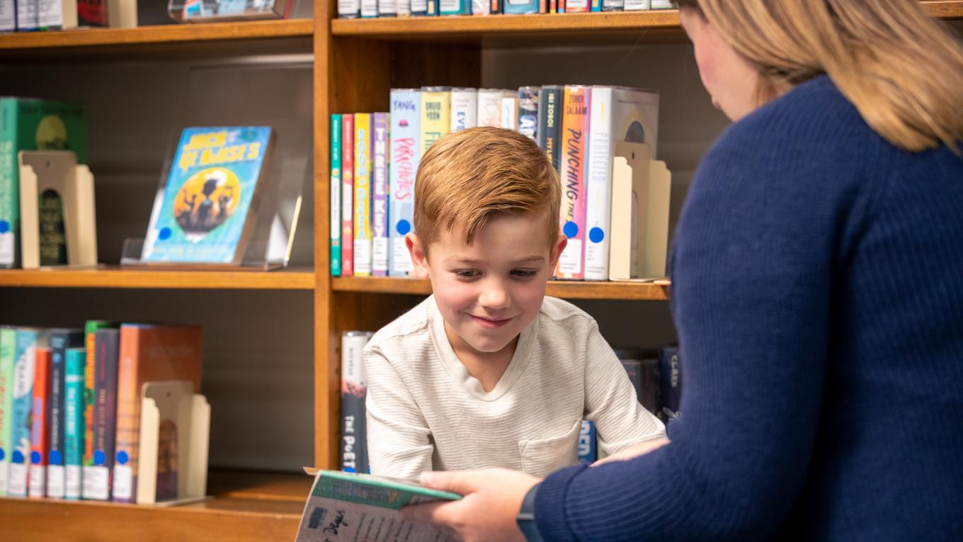 Young learner smiling and looking at a book in the hands of Duquesne Reading Specialist Certification student and both are sitting in front of library bookshelves.