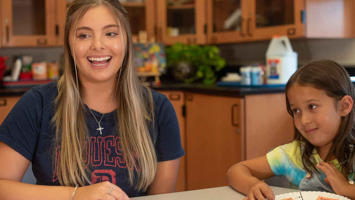  Duquesne student teacher engaging in lesson at table with K-12 student next to her in clasroom with paper plate science project