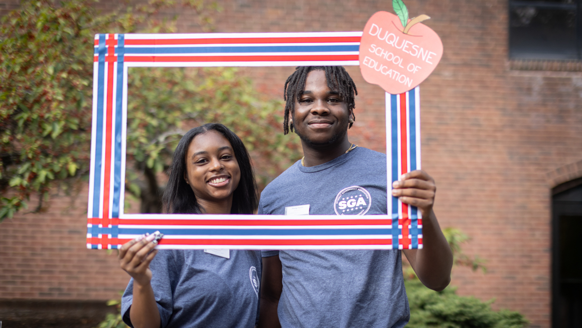 School of Education's Jayla Flenory with colleague smiling as framed by a Duquesne School of Education photo prop frame outside Canevin Hall on campus.