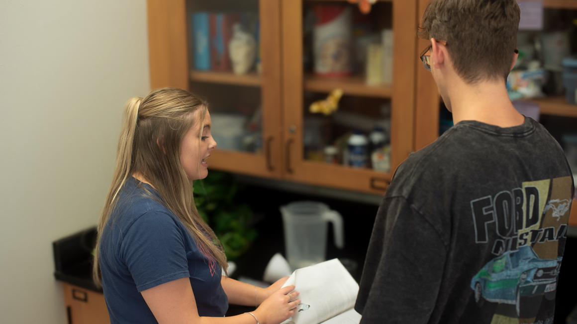 Student teacher in classroom reading science book standing with high school student