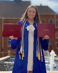 School of Education B.S.Ed. Secondary Education alumni in graduation attire holding two diplomas smiling in front of fountain on campus on a blue sky day