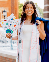 B.S.Ed. Early Childhood Education PreK-4 alum Julia Arlia in graduate attire outside on campus in front of a fountain holding cap that reads the world is my classroom.