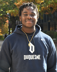 B.S.Ed. Secondary Education student Mwango Kasuba wearing a football sweatshirt and necklace outside on campus with autumn trees in the background.