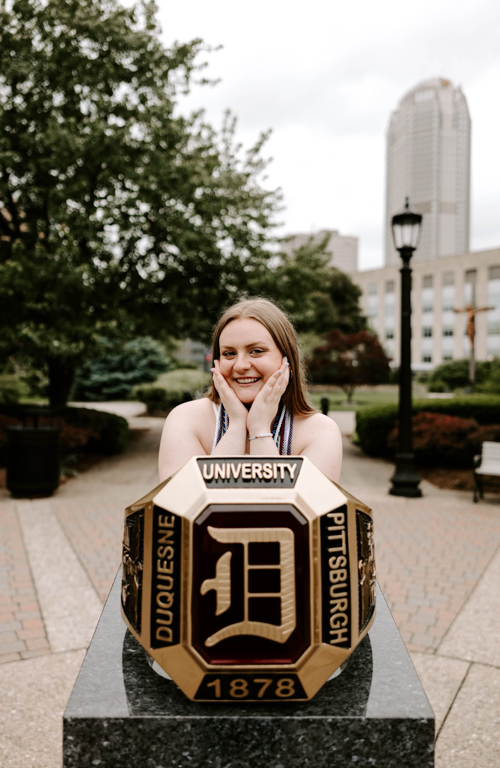 B.S.Ed. Early Childhood Education alum Alyssa Napolitana behind ring monument posed with eblows on monument and hands framing face outside on campus with green trees and city buildings in background