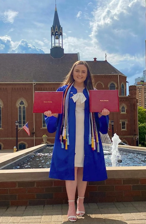 School of Education B.S.Ed. Secondary Education alumni in graduation attire holding two diplomas smiling in front of fountain on campus on a blue sky day