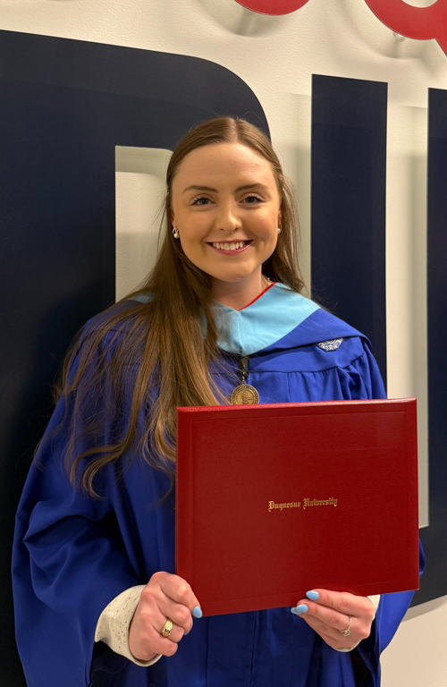 Photo of M.S.Ed. Education Administration graduate Amber DiMaria in graduate regalia holding diploma smiling on commencement day with walll with Duquesne colors and parts of letter behind her