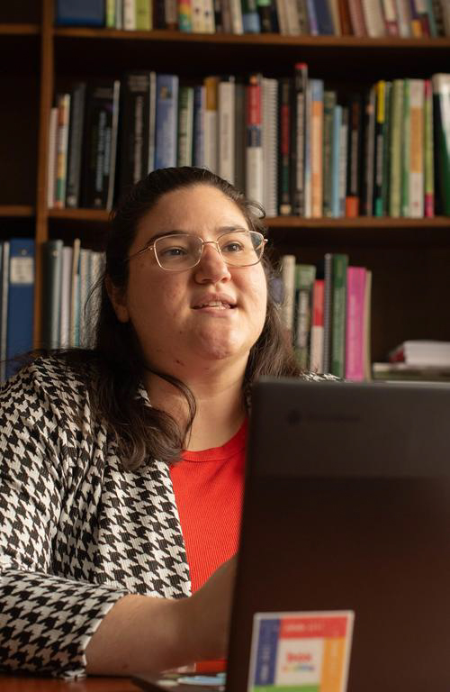 Ph.D. School Psychology graduate student Athena Vafiadis engaged in work looking ahead in discussion with laptop in front of her and bookshelves in the background.