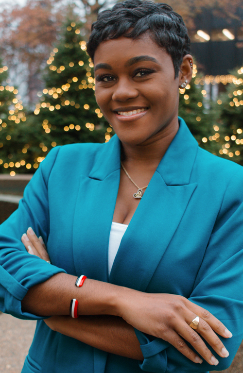 Fulbright Scholar and Ph.D. School Psychology student outside union smiling with arms crossed wearing blazer and Christmas trees with lights in background