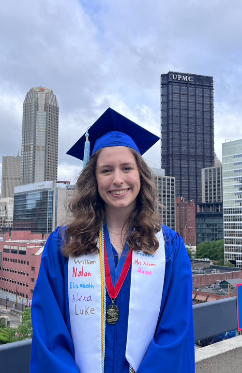 B.S.Ed. Early Childhood Education alum Isabel Rothstein in graduation regalia outside with cityscape background from Duquesne Univesity's campus behind her