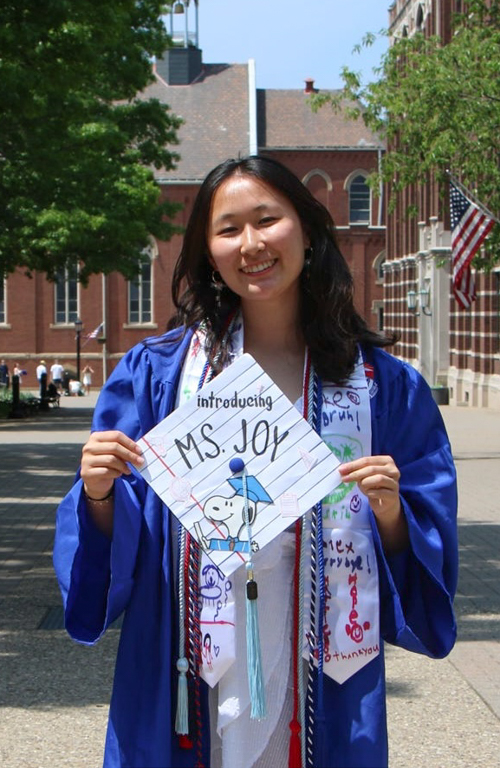 Fulbright Scholar and B.S.Ed. Early Childhood alum Joy Chen standing on Duquesne's campus in graduation regalia smiling and holding decorated graduation cap that reads Introducing Ms. Joy with Snoopy in grad cap holding diploma.