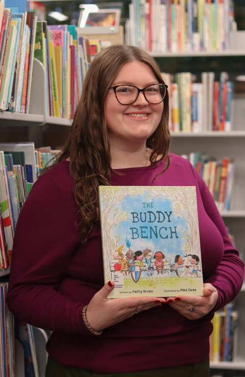 School of Education B.S.Ed. Dual Certification student Kendall House smiling with children's Buddy Bench book in hand against backdrop of colorful library bookshelves.