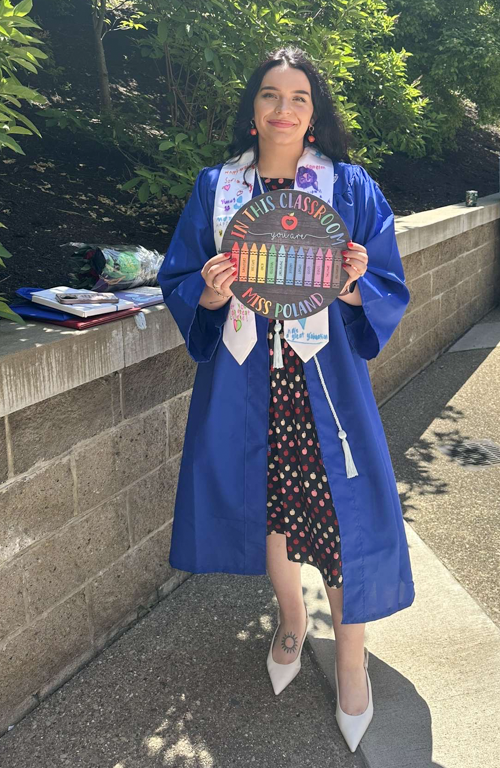 School of education alum and student Kylie Poland in graduation regalia smiling outside holding sign with colored crayons that reads