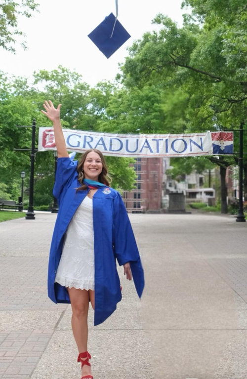 MAT Early Childhood education alum Madison Dayton in graduate regalia on campus throwing graduation ap in air as she smiles looking straight ahead and the Graduation banner framing her in background over academic walk.