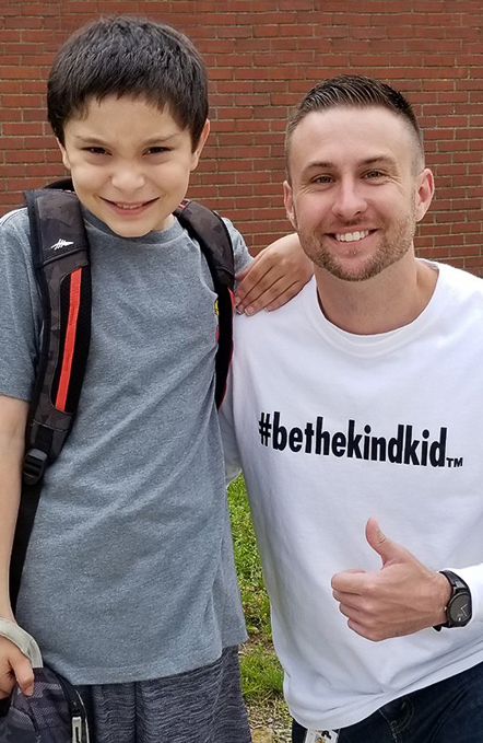 Photo of Ed.D. Educational Leadership graduate student and Hosack Elementary Principal Matt Heckmann wearing shirt that reads #BeTheKindKid as he smiling next to elementary school student outside of the school