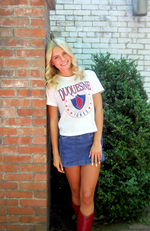 Early Education major Molly Manzer standing by a brick wall in a Duquesne t-shirt and jean skirt.