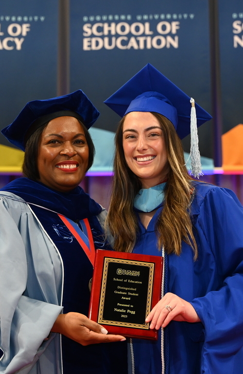 Natalie Pegg with graduate award in hand smiling on stage next to School of Education Dean Generett.