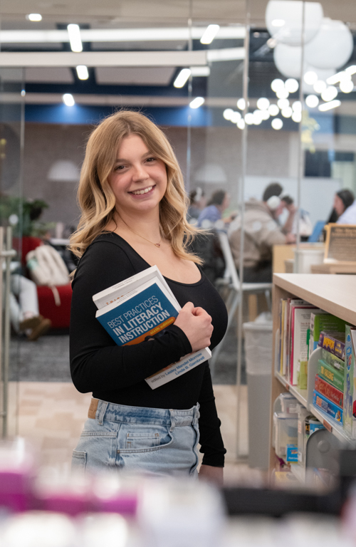 School of Education undergraduate student Riley Adams holding book in library and smiling.