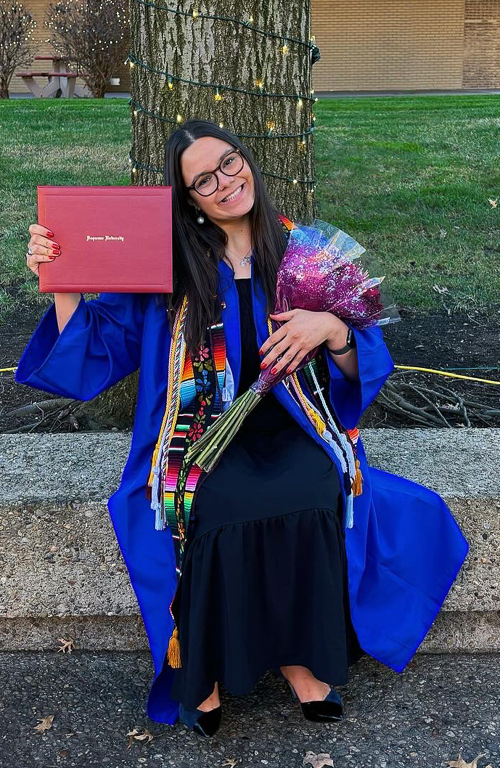 Sarah Lopez sitting on campus wearing graduation regalia holding diploma and flowers in hands smiling