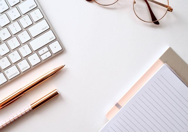 A computer keyboard, two pens, eyeglasses, and a small notebook lay ontop of a white surface.